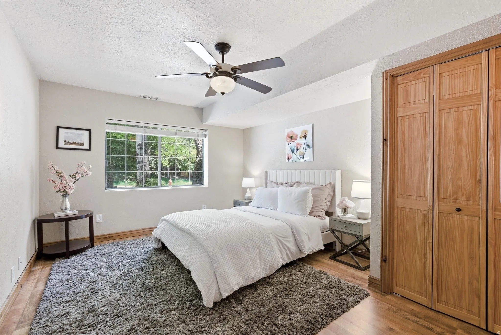 Bedroom with wood-finish flooring, a ceiling fan with integrated lighting, and a large window overlooking trees