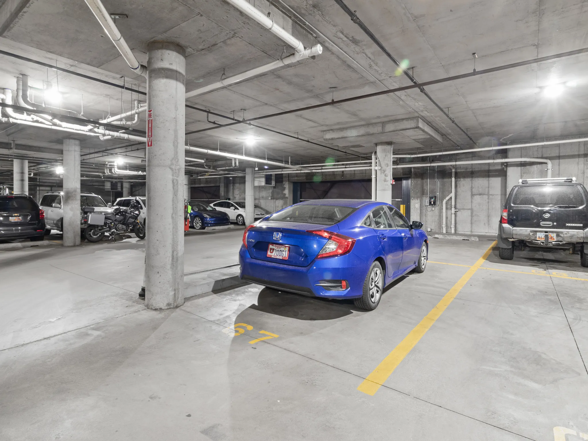 Concrete garage with overhead exposed piping, illuminated by multiple ceiling-mounted light fixtures
