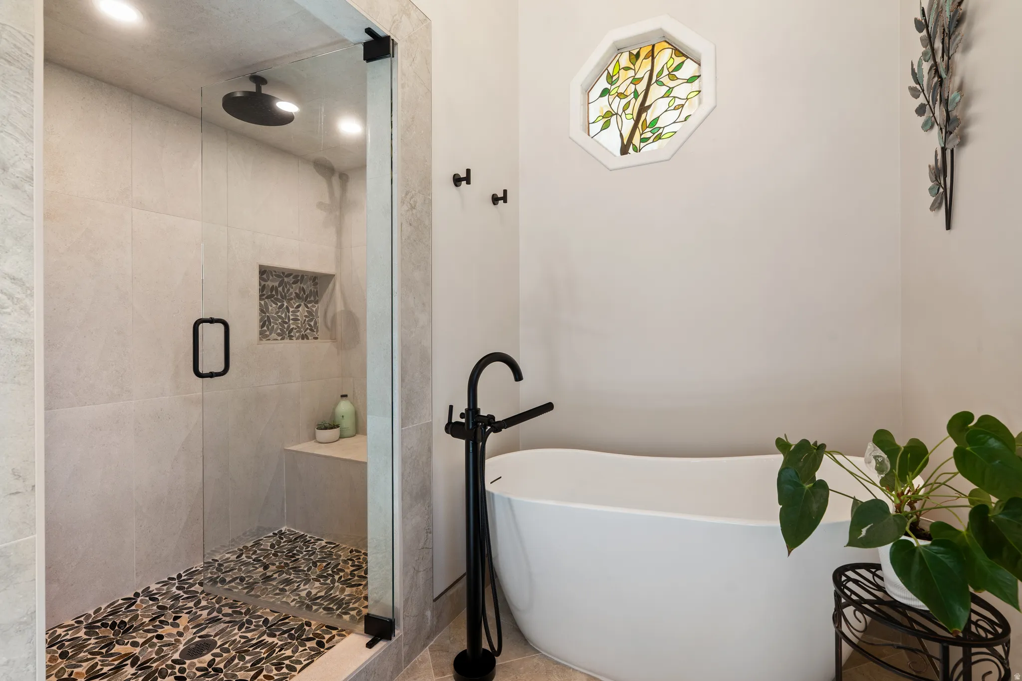 Freestanding soaking tub with a floor-mounted matte black faucet, walk-in shower with a frameless glass door, pebble floor, built-in bench, and a unique stained-glass octagonal window