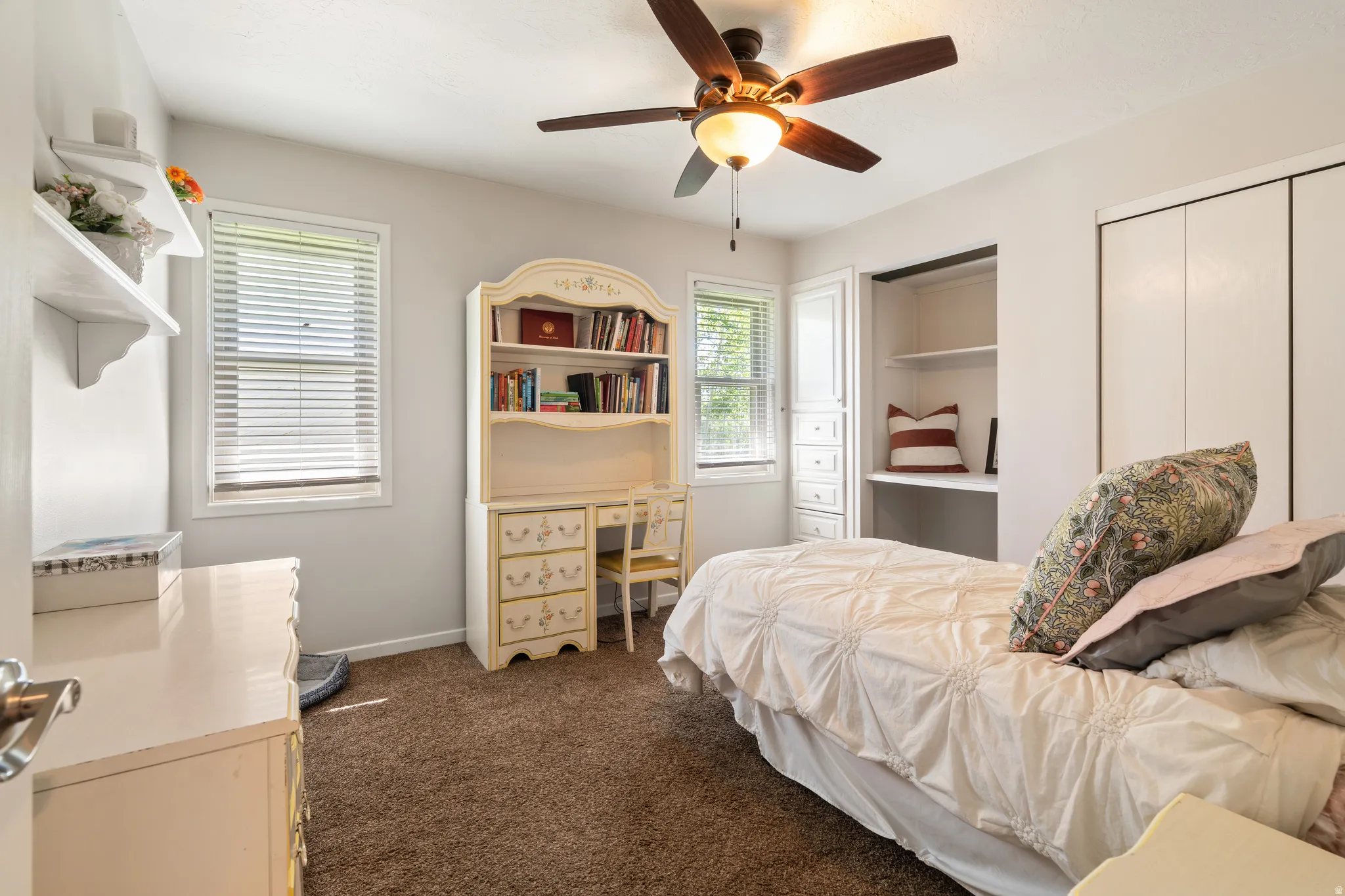 Carpeted room with a ceiling fan, two windows with blinds, a built-in desk with shelving, and a built-in closet with bifold doors
