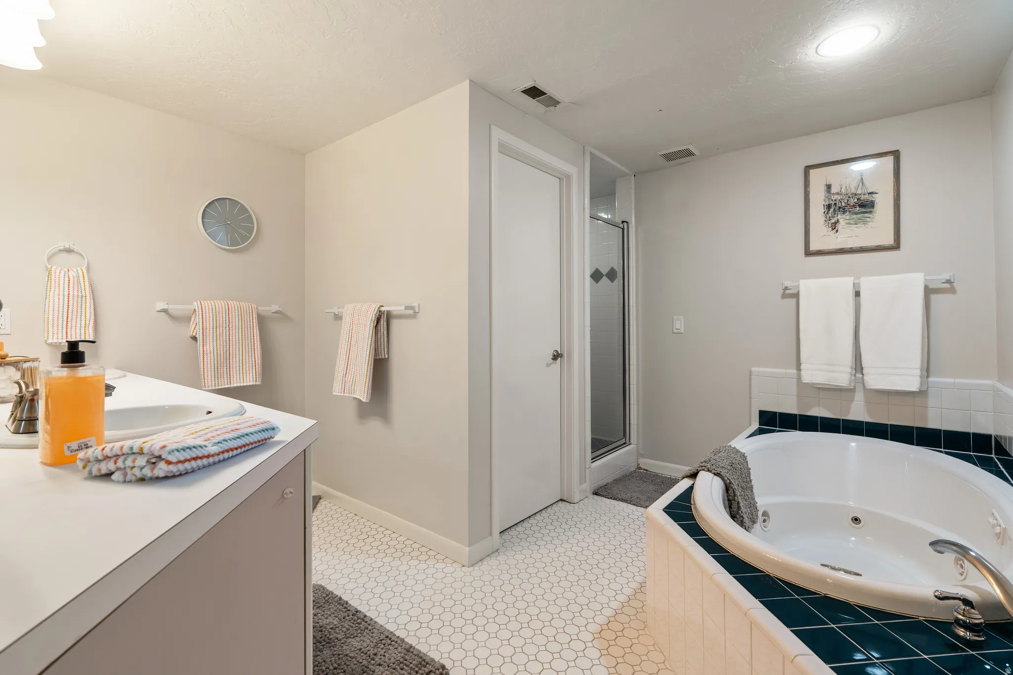 Bathroom featuring a corner jetted tub with tile surround, a separate glass-enclosed shower, a vanity with a white countertop and integrated sink, and white hexagonal tile flooring