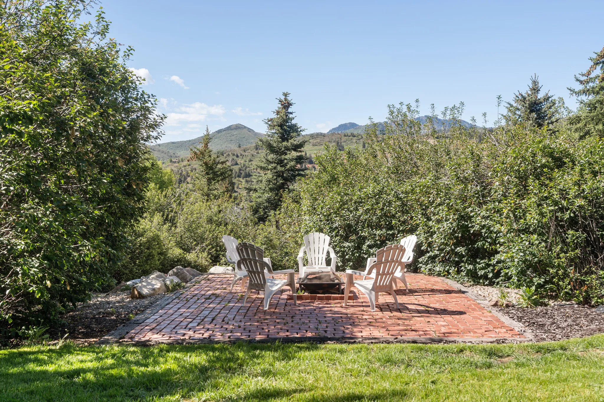 Brick paver patio with a built-in fire pit, surrounded by lush green foliage and mature trees, featuring a distant mountain backdrop