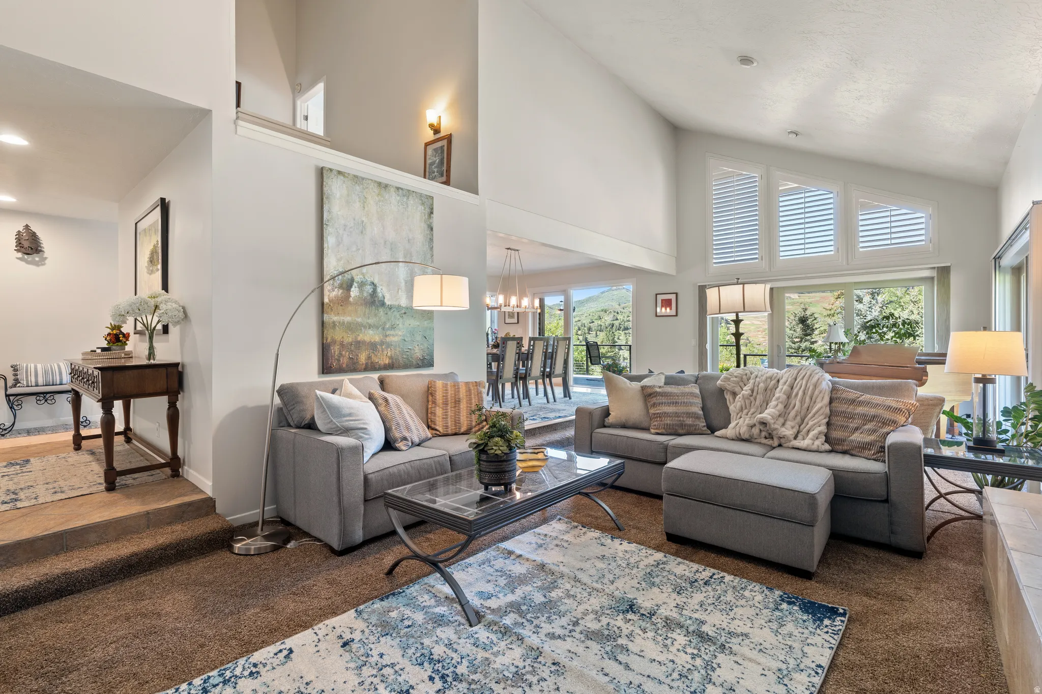 Open-concept living space featuring vaulted ceilings, a prominent window wall with custom shutters, and plush carpeting