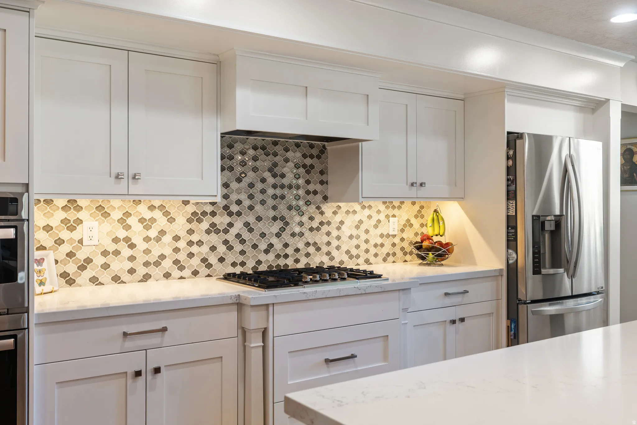 Kitchen featuring white shaker cabinetry, patterned tile backsplash, gas cooktop, under-cabinet lighting, and stainless steel refrigerator