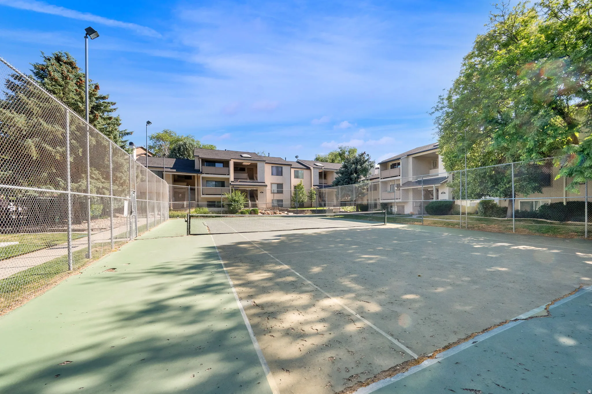 Tennis court with chain-link fencing, surrounded by mature trees and multi-level residential buildings