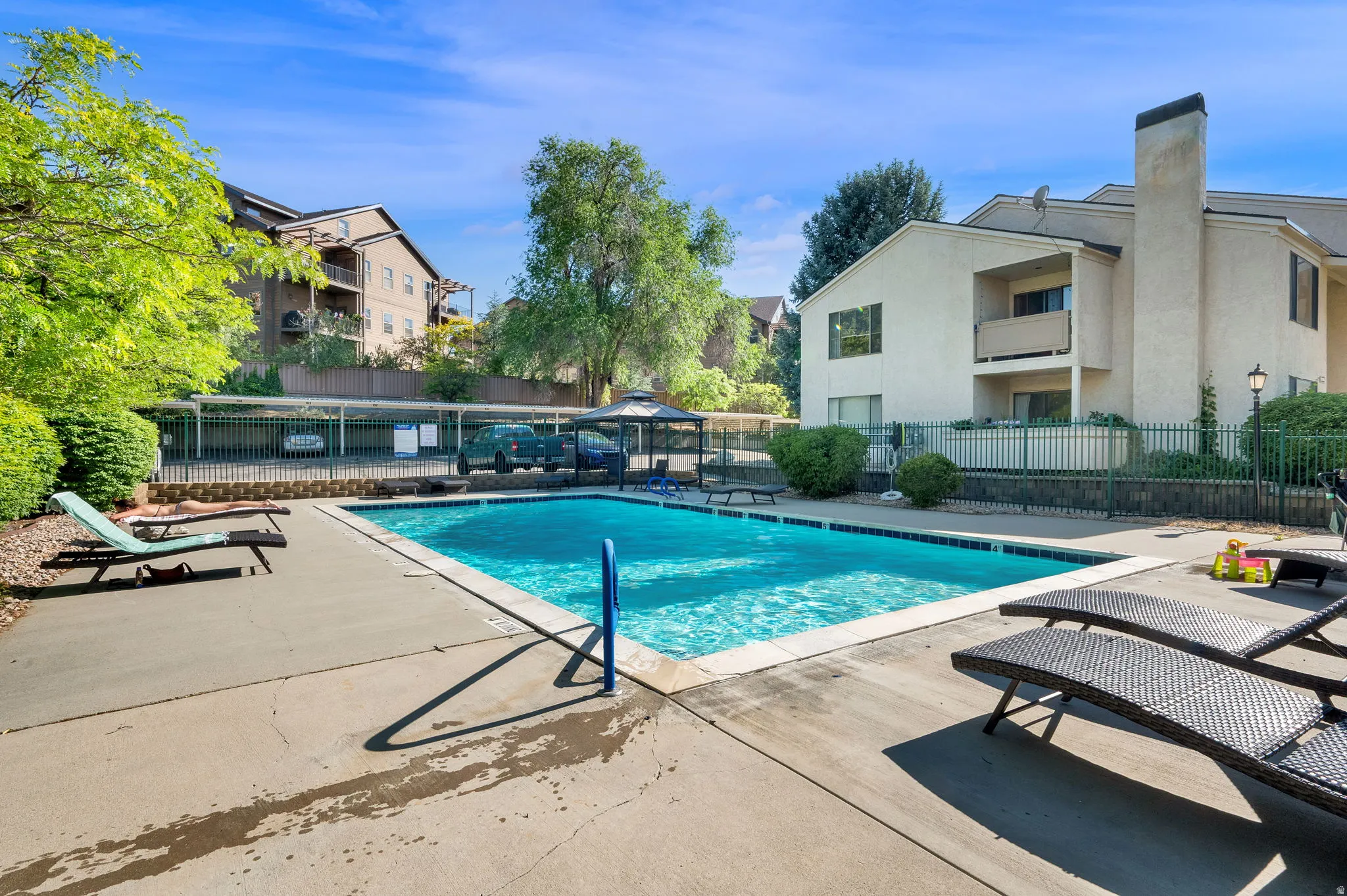 Outdoor swimming pool with concrete decking, surrounded by mature landscaping and a black metal fence