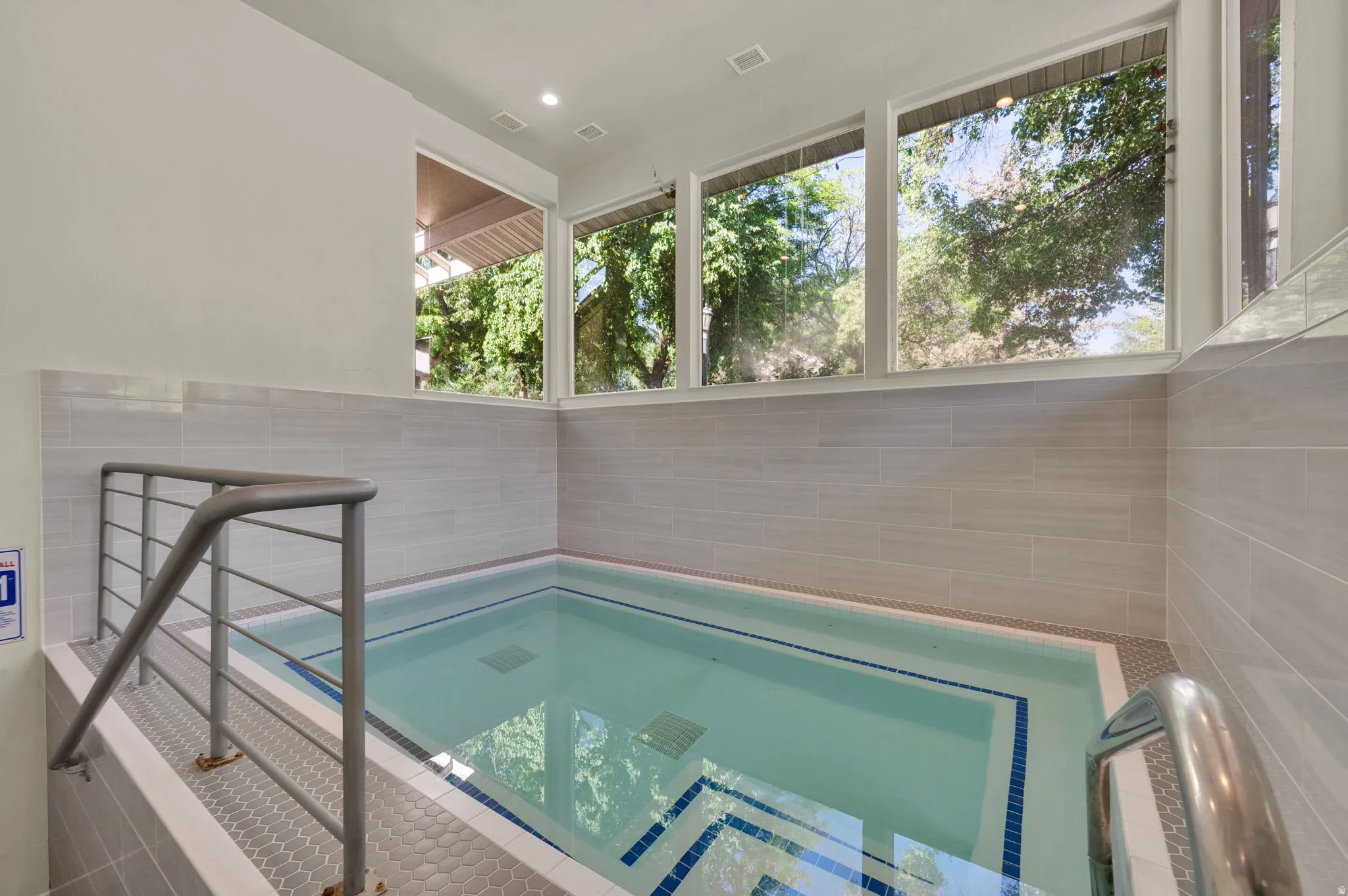 Indoor spa pool featuring a metal handrail, light gray wall tiling, and a series of upper windows