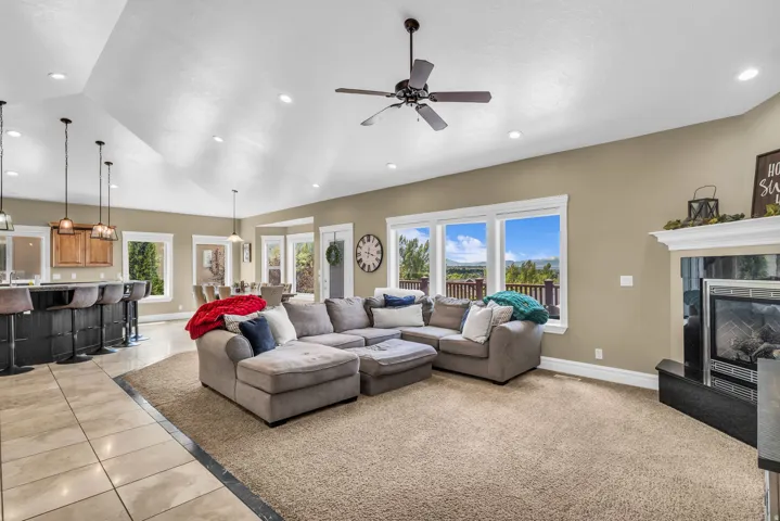 Open-concept living area featuring vaulted ceilings, recessed lighting, a ceiling fan, a built-in fireplace with a mantel, and multiple windows offering expansive outdoor scenery