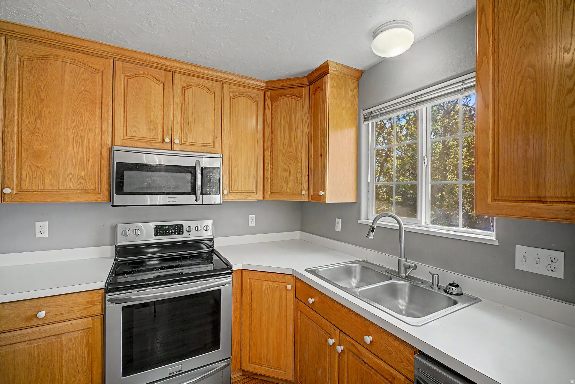 Kitchen featuring wood cabinetry, stainless steel appliances, white countertops, and a dual-basin stainless steel sink with a window overhead