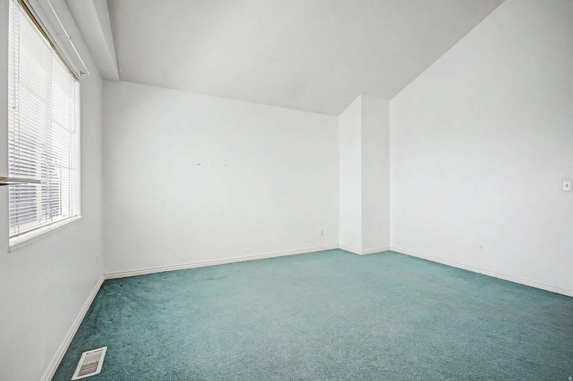 Spacious interior room featuring a large window with horizontal blinds, light-colored walls, and green carpet flooring