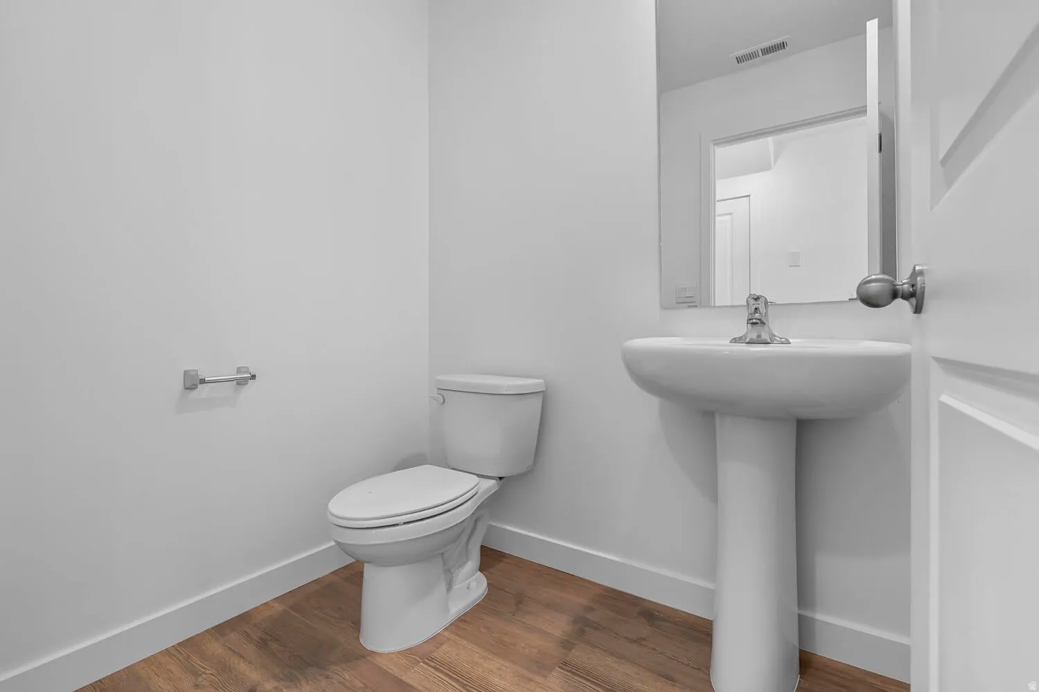 Modern powder room featuring a pedestal sink with chrome faucet, a white toilet, wood-finish flooring, and a wall-mounted mirror