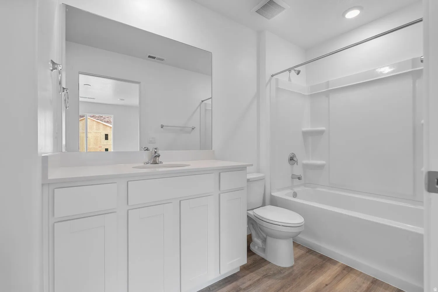 Bathroom featuring a white vanity with integrated sink, a large wall-mounted mirror, a tub/shower combination with white surround, a toilet, and wood-finish flooring