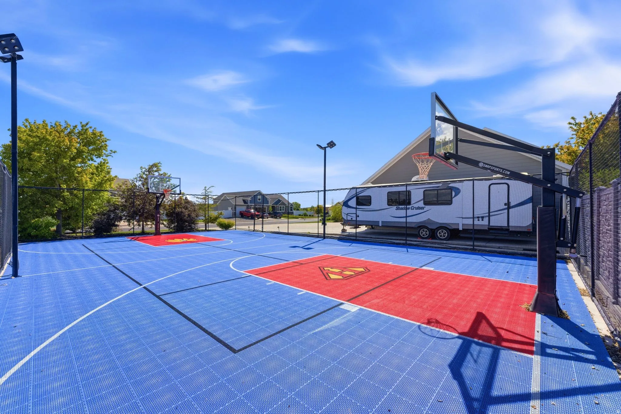 Multi-sport court featuring blue and red interlocking tile surfacing, two basketball hoops, perimeter fencing, and an integrated tall light fixture