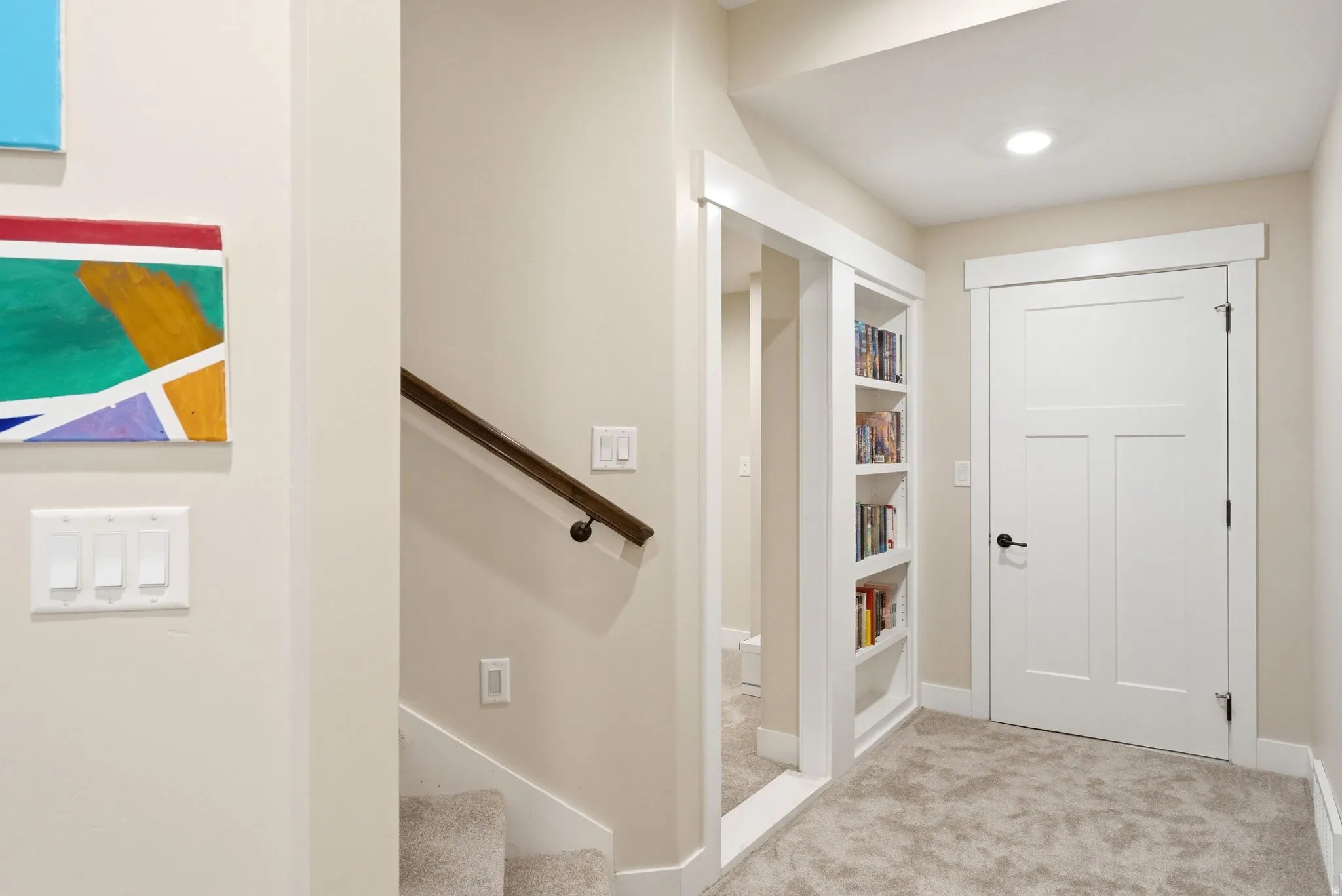 Carpeted hallway featuring a built-in bookshelf, a white paneled door, and a staircase with a wooden handrail
