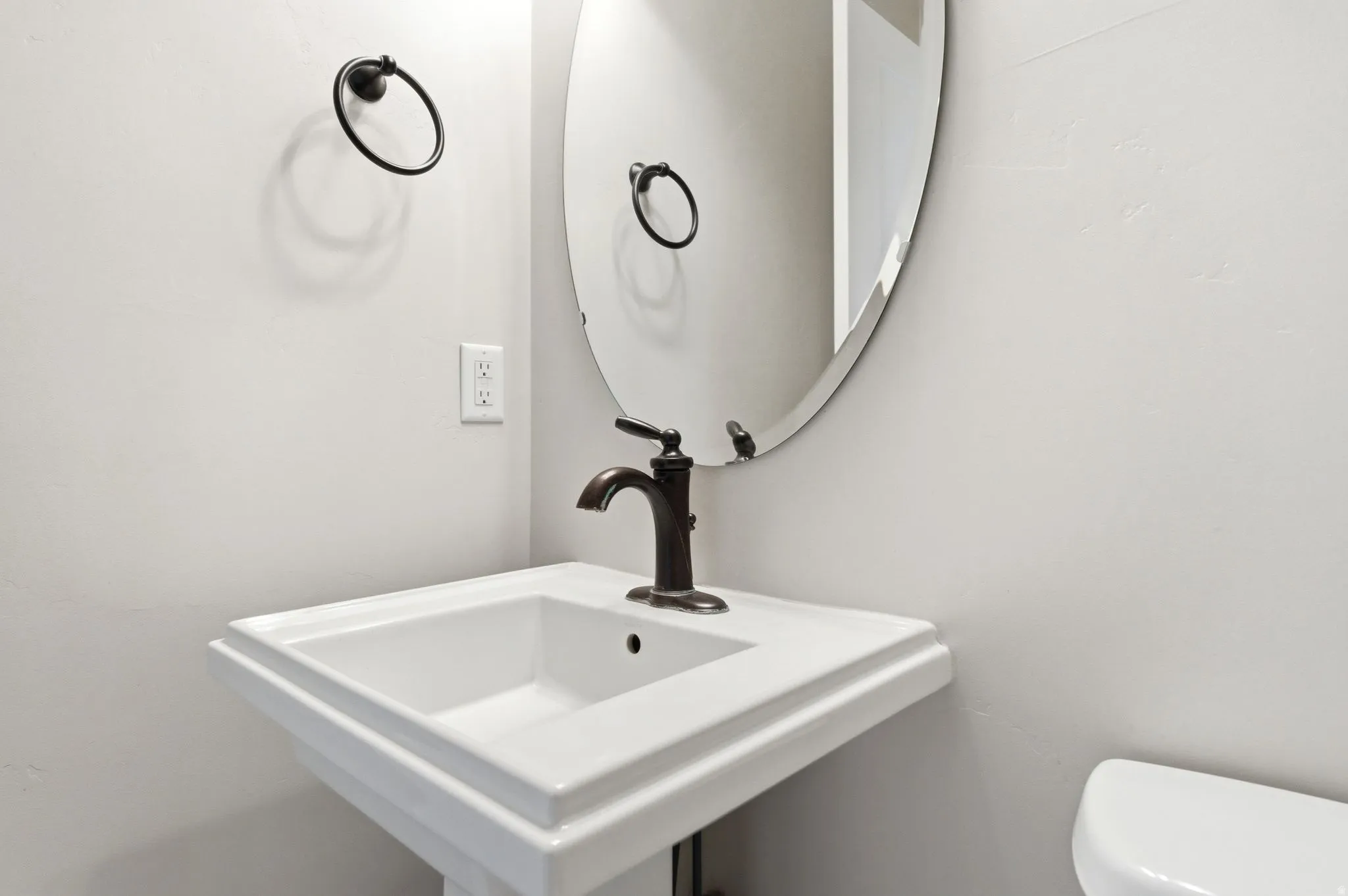 White pedestal sink with a bronze-finish faucet, accompanied by an oval frameless mirror and a coordinating bronze-finish towel ring