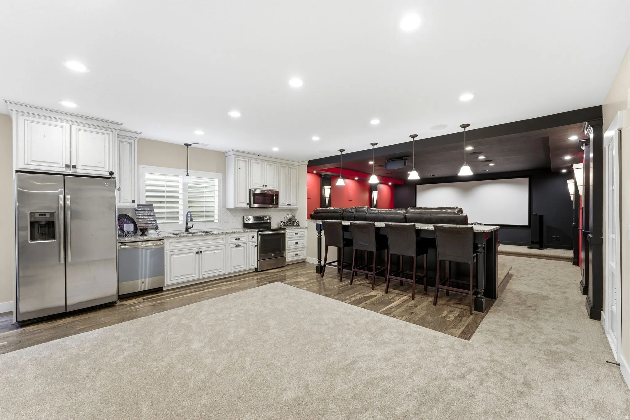 Spacious entertainment area featuring a full kitchen with white cabinetry and stainless steel appliances, a multi-stool bar island, a large projection screen, and recessed lighting throughout