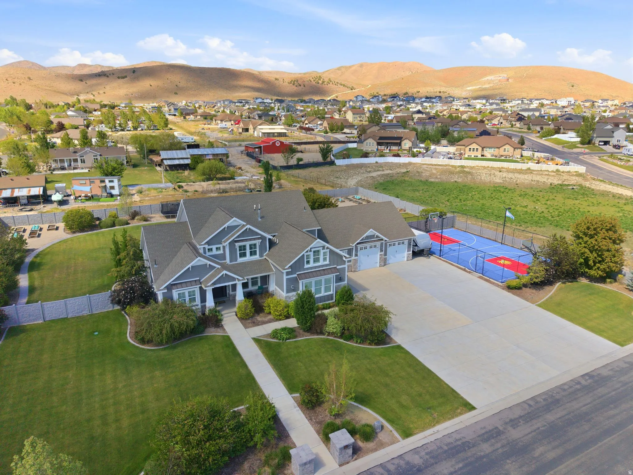 Spacious property featuring a large residence with gray siding and white trim, an attached multi-car garage, an expansive concrete driveway, a dedicated sports court, and manicured lawns with established landscaping