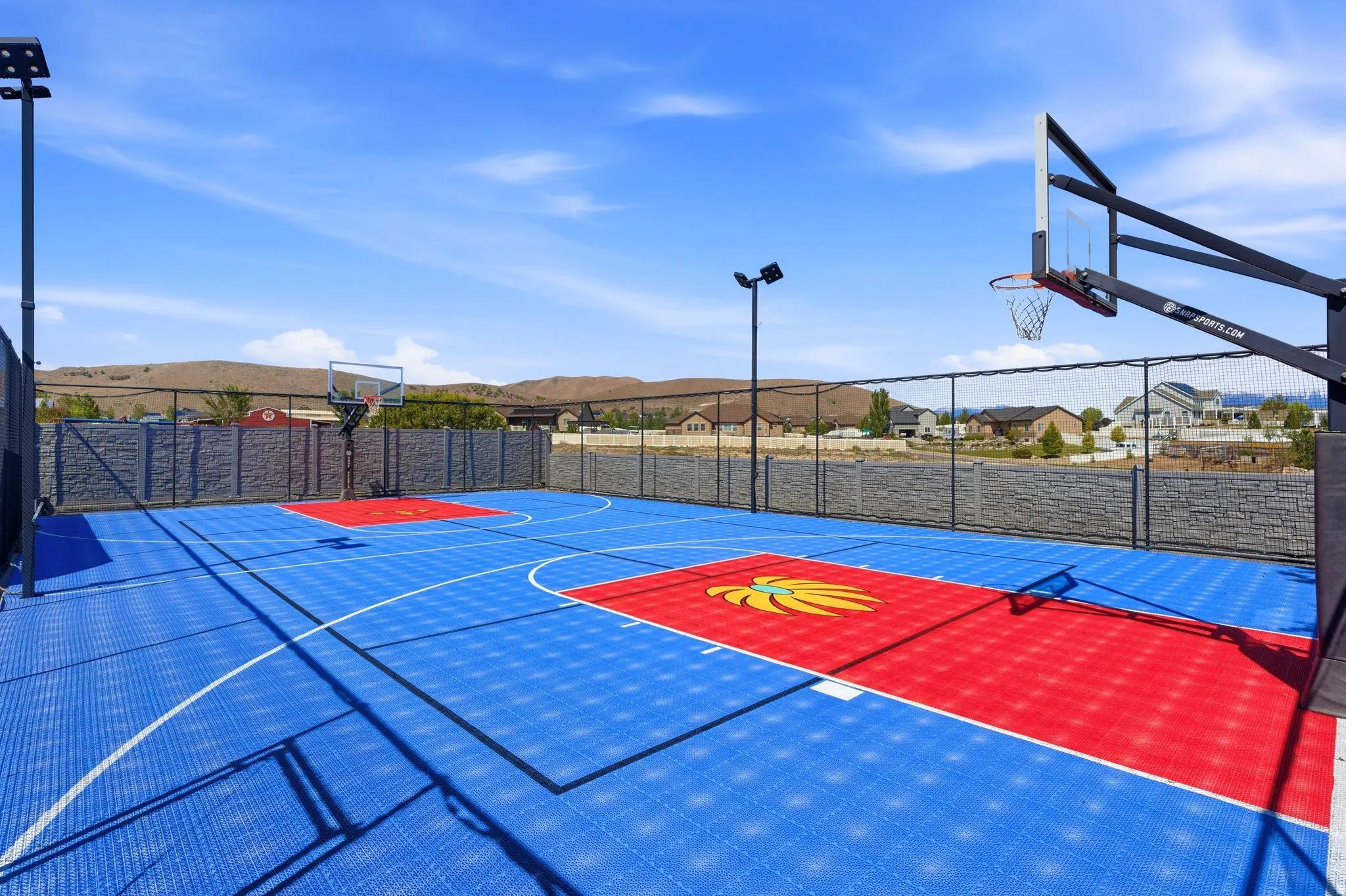 Multi-sport court featuring blue and red surface tiling, two basketball hoops, perimeter fencing, and integrated sports lighting