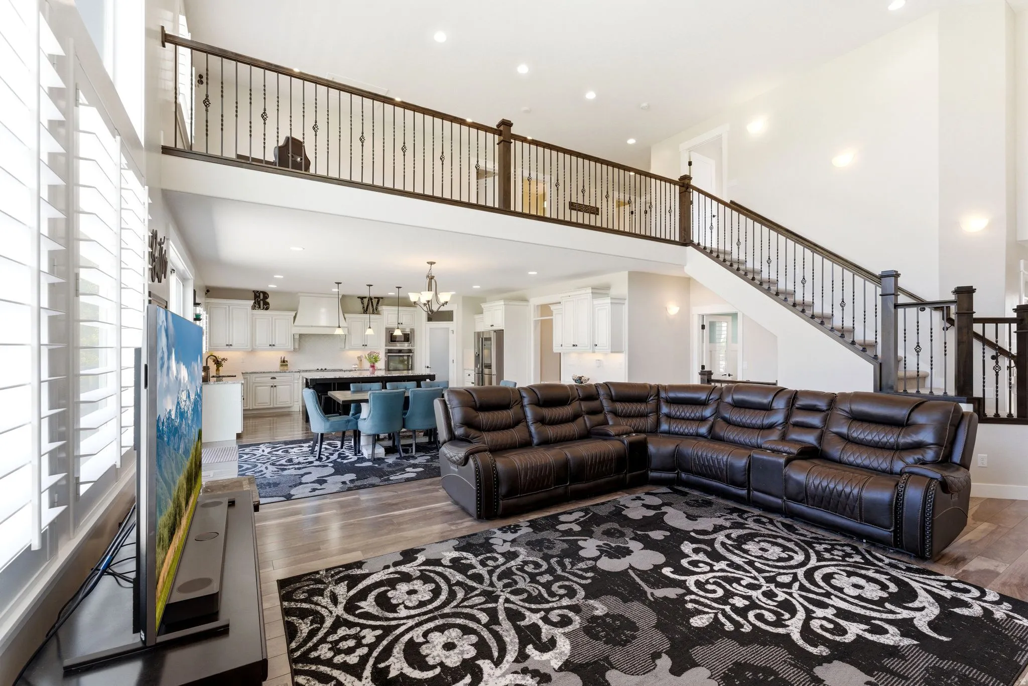 Open-concept living area featuring a two-story ceiling, hardwood-style flooring, a staircase with dark wood banisters and wrought iron balusters, and a kitchen with white cabinetry and a dark-toned island