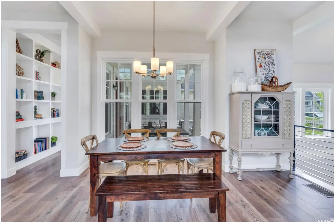 Open-concept dining area featuring wood-finish flooring, a contemporary chandelier, multiple windows with white trim, a built-in shelving unit, and a railing with horizontal metal spindles