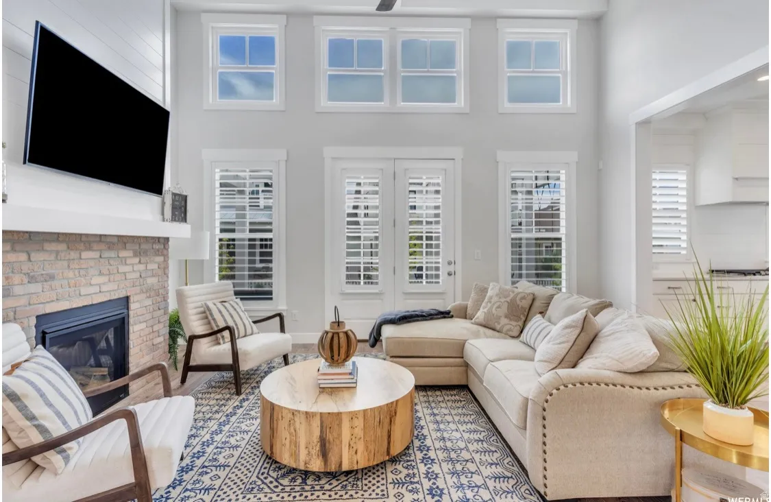Two-story living area featuring a stacked stone fireplace with a white mantel, multiple windows with white plantation shutters, and a wood-finish flooring with a patterned area rug