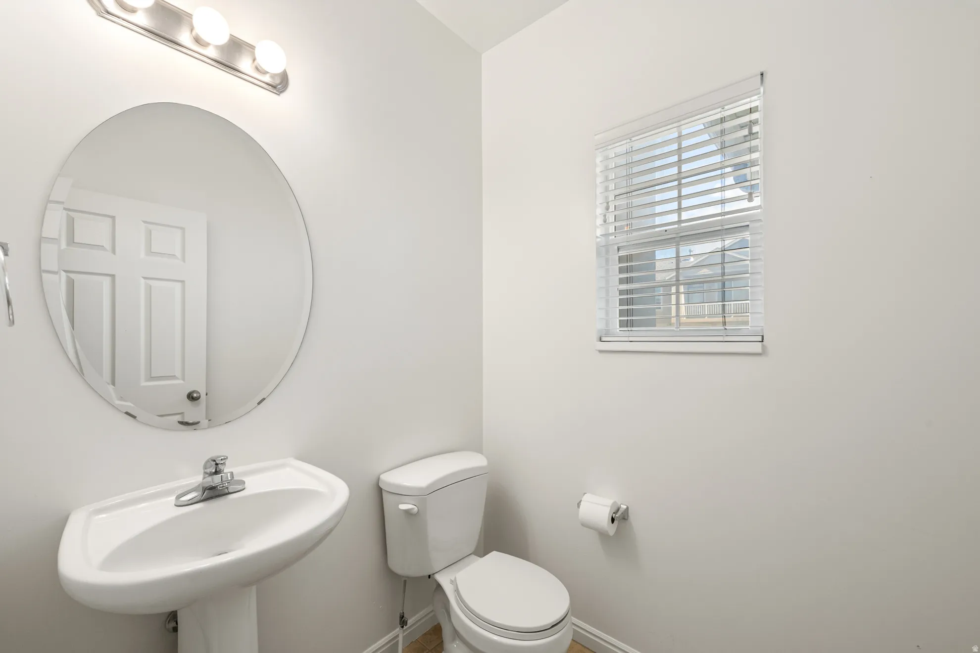 Pedestal sink with chrome faucet, complemented by a round mirror and a three-light vanity fixture