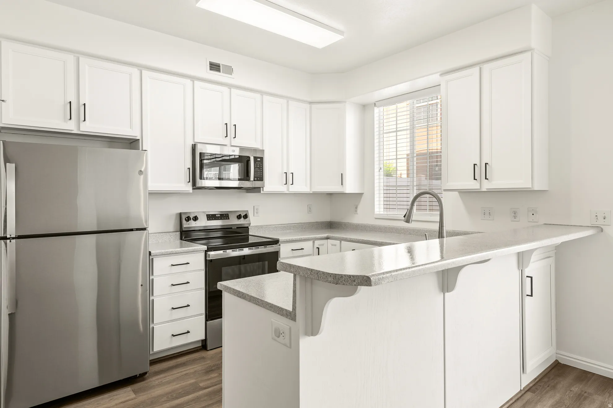 Bright kitchen featuring white cabinetry, stainless steel appliances, speckled countertops, wood-finish flooring, and a breakfast bar