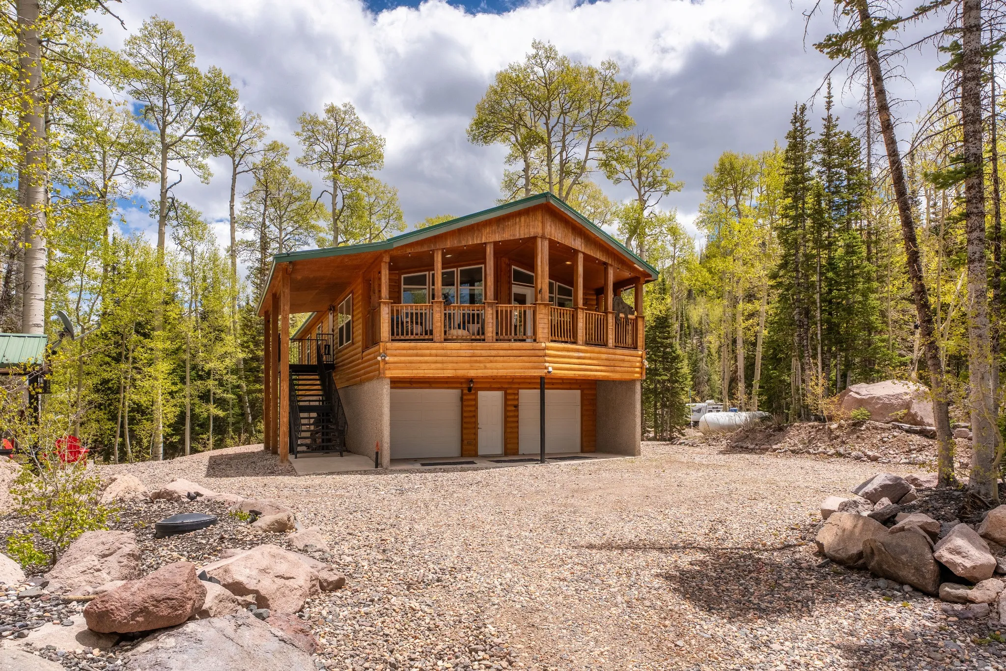 Wood-sided cabin featuring a covered balcony, integrated garage, exterior staircase, and gravel driveway, surrounded by mature trees