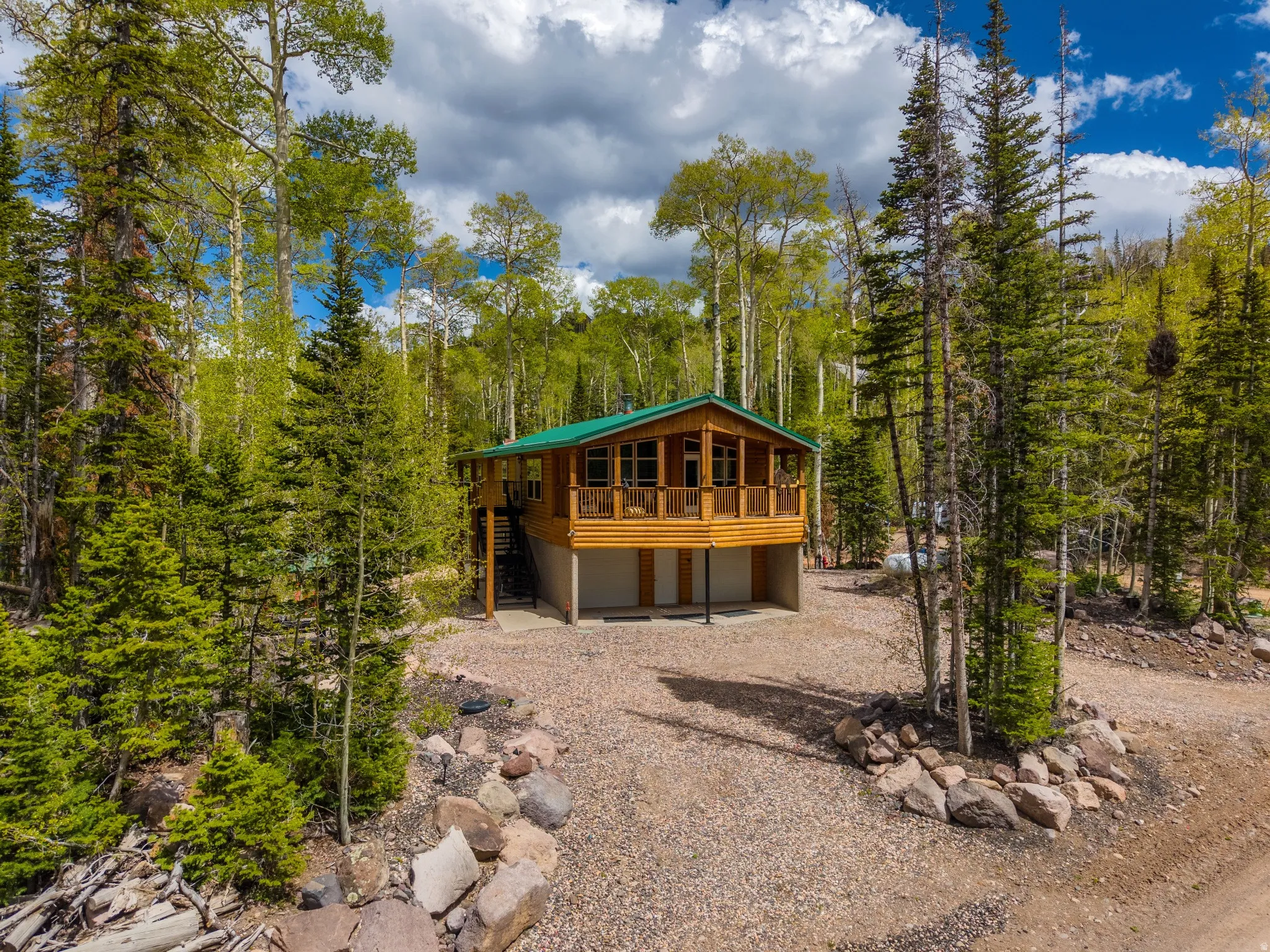 Log-sided cabin with a green metal roof, elevated on concrete stilts, featuring a wraparound wood-plank deck with a railing, and a gravel driveway surrounded by mature trees