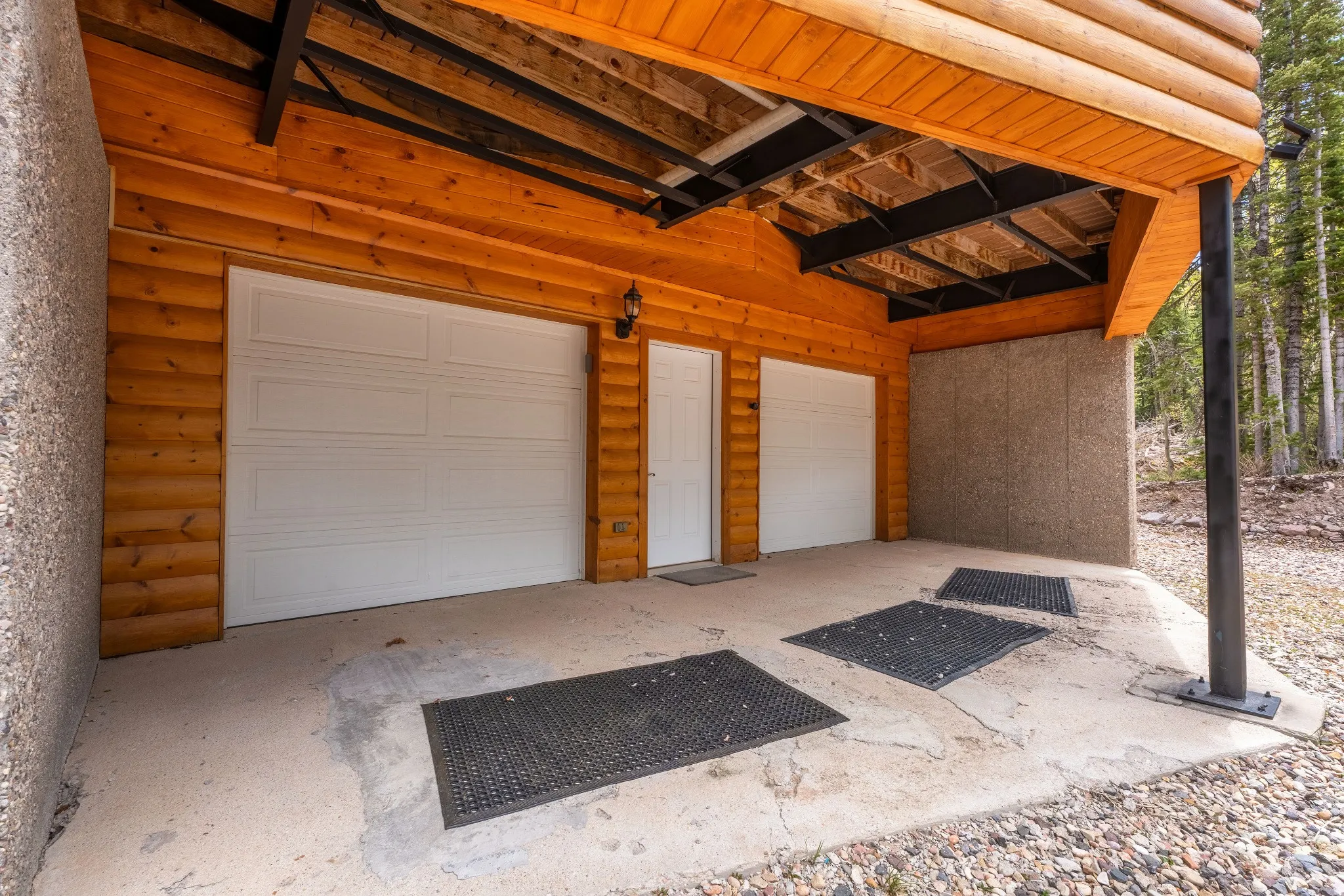 Two white garage doors with overhead lighting, wood-paneled exterior, exposed wooden ceiling beams, concrete pad, and a forest backdrop