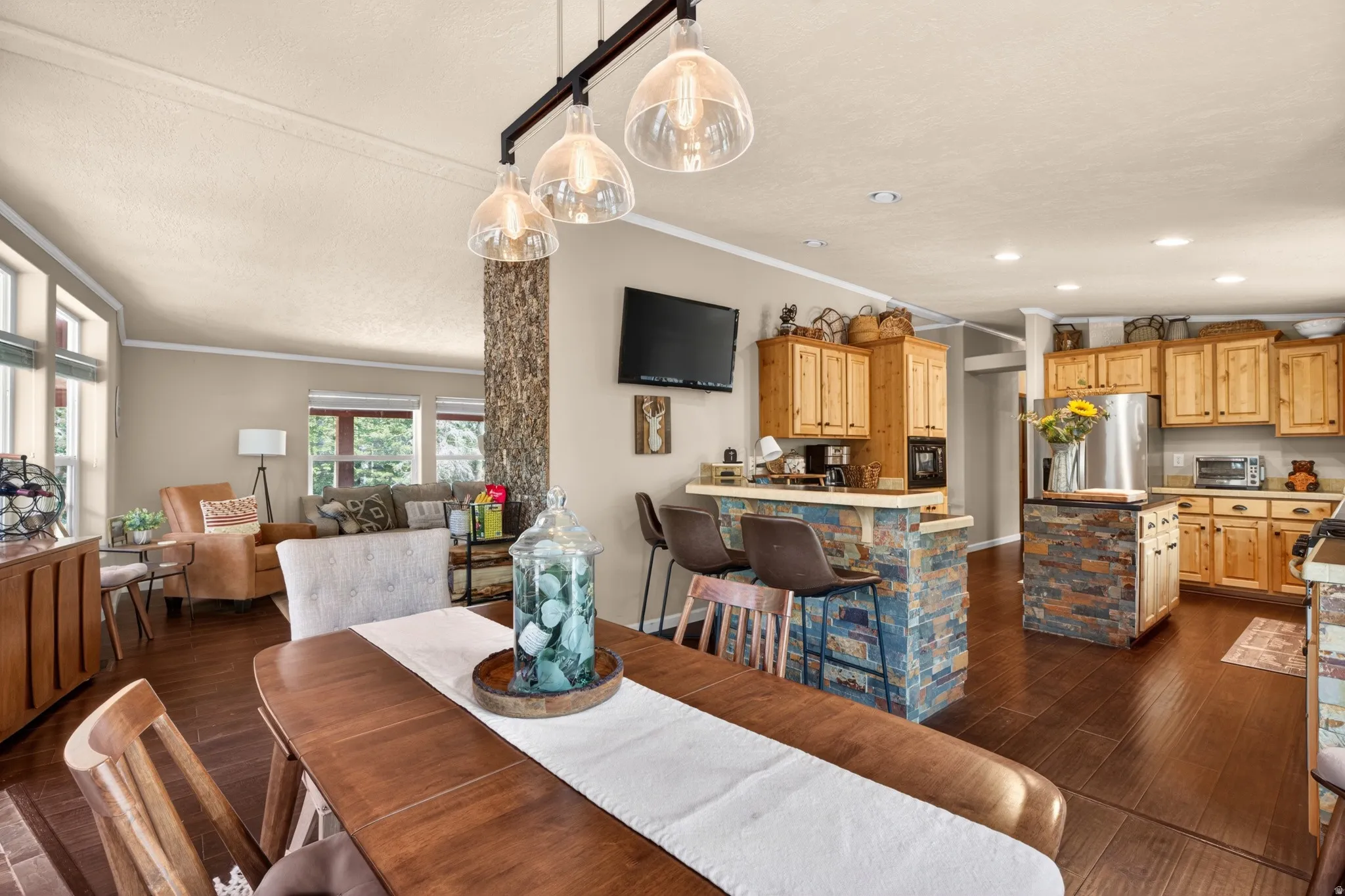 Open-concept living space featuring wood-finish flooring, a dining area with a linear pendant light fixture, a kitchen with light wood cabinetry, a stone-clad kitchen island, and a breakfast bar with stone accents