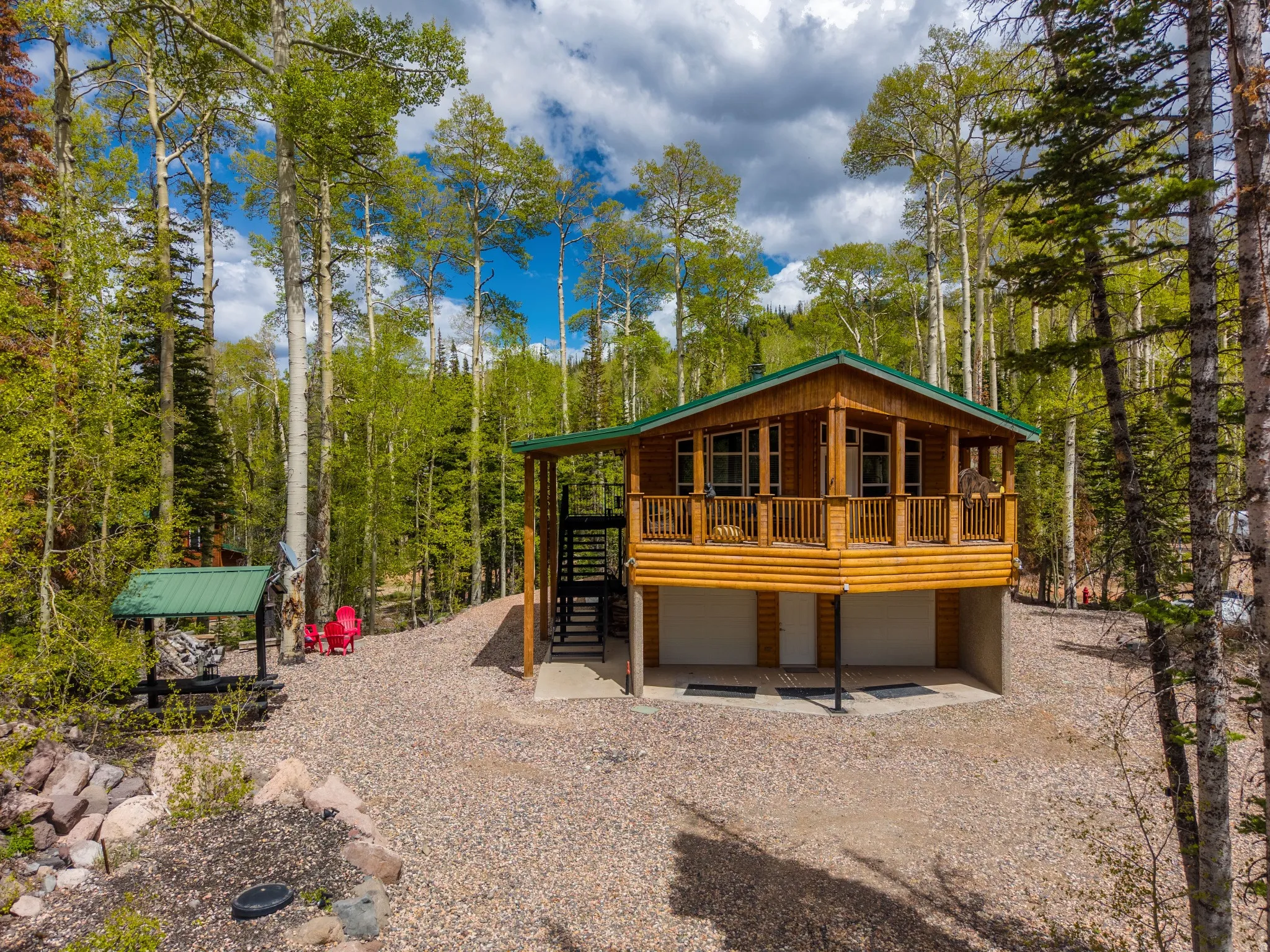 Log cabin exterior featuring a green metal roof, a covered porch with wood railing, an exterior staircase, and a gravel driveway