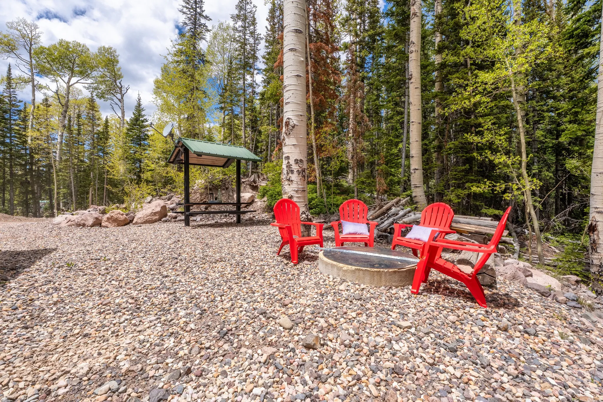 Gravel outdoor area with a built-in fire pit, surrounded by mature trees, and featuring a covered shelter