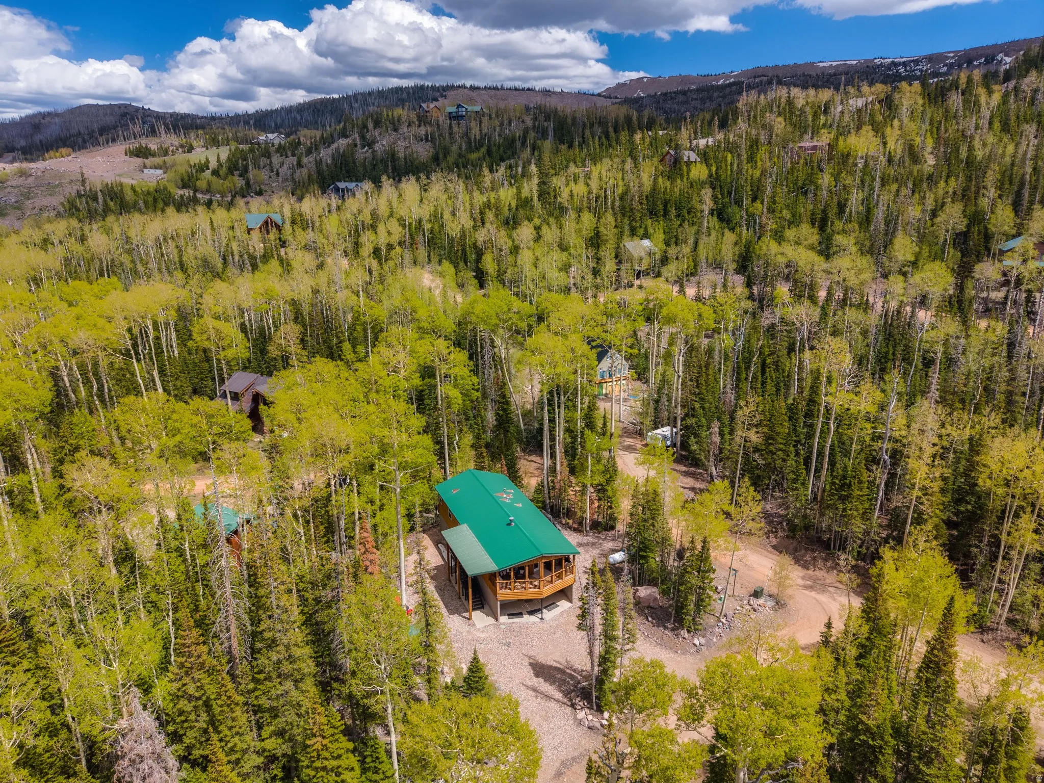 Wooded mountain property featuring a cabin with a green roof, a wraparound deck, and a gravel driveway