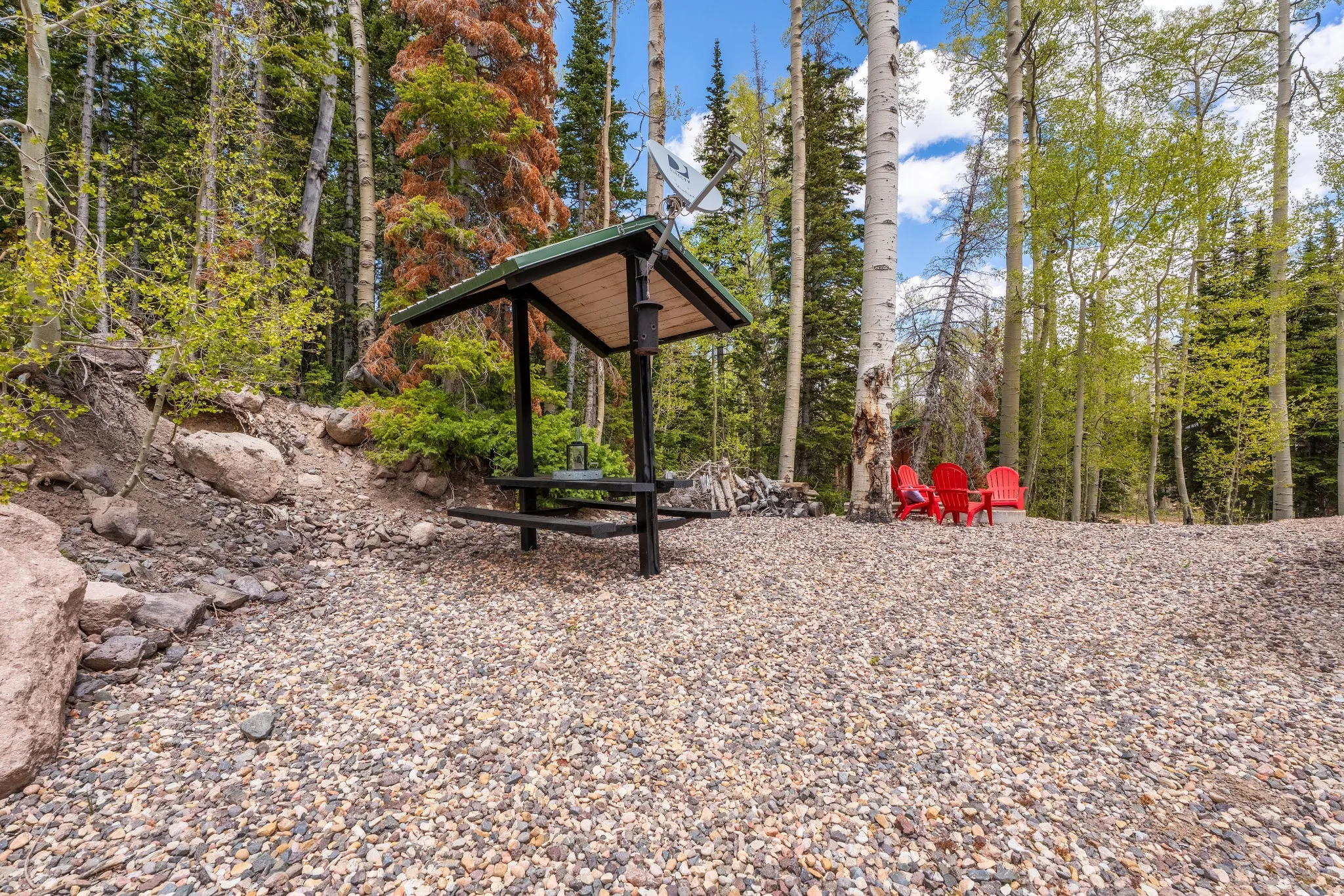 Gravel outdoor area featuring a covered picnic structure with an integrated grill, surrounded by mature trees and natural rock formations