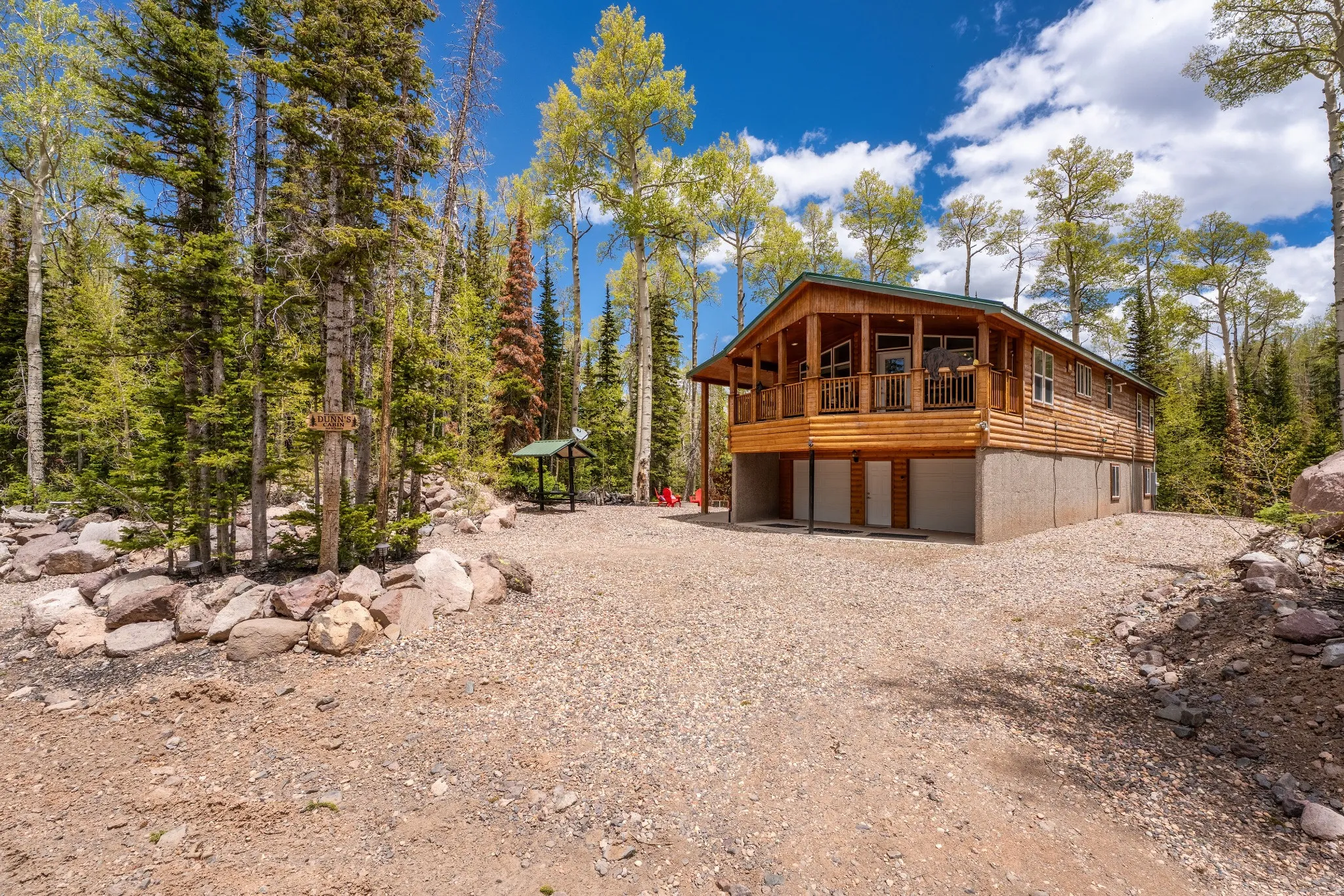Log-sided cabin surrounded by mature trees, featuring a covered porch with wooden railings and a gravel driveway