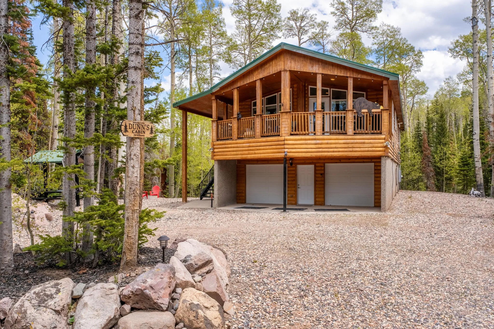 Log-sided residence featuring an elevated covered porch with a wood railing, a gravel driveway, and two garage bays