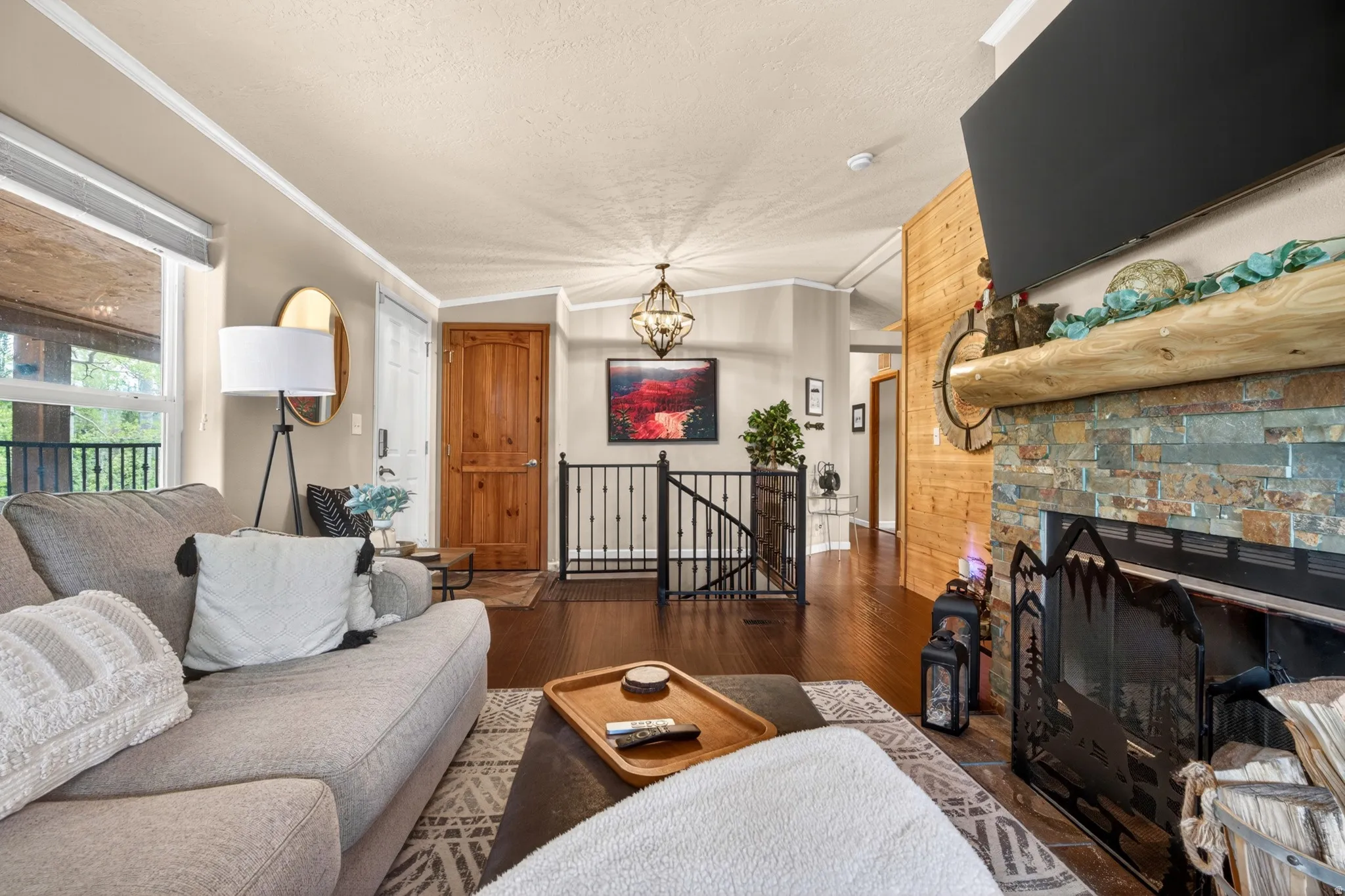Living area featuring a stone-clad fireplace with a prominent log mantel, wood-finish flooring, and a large window overlooking a balcony