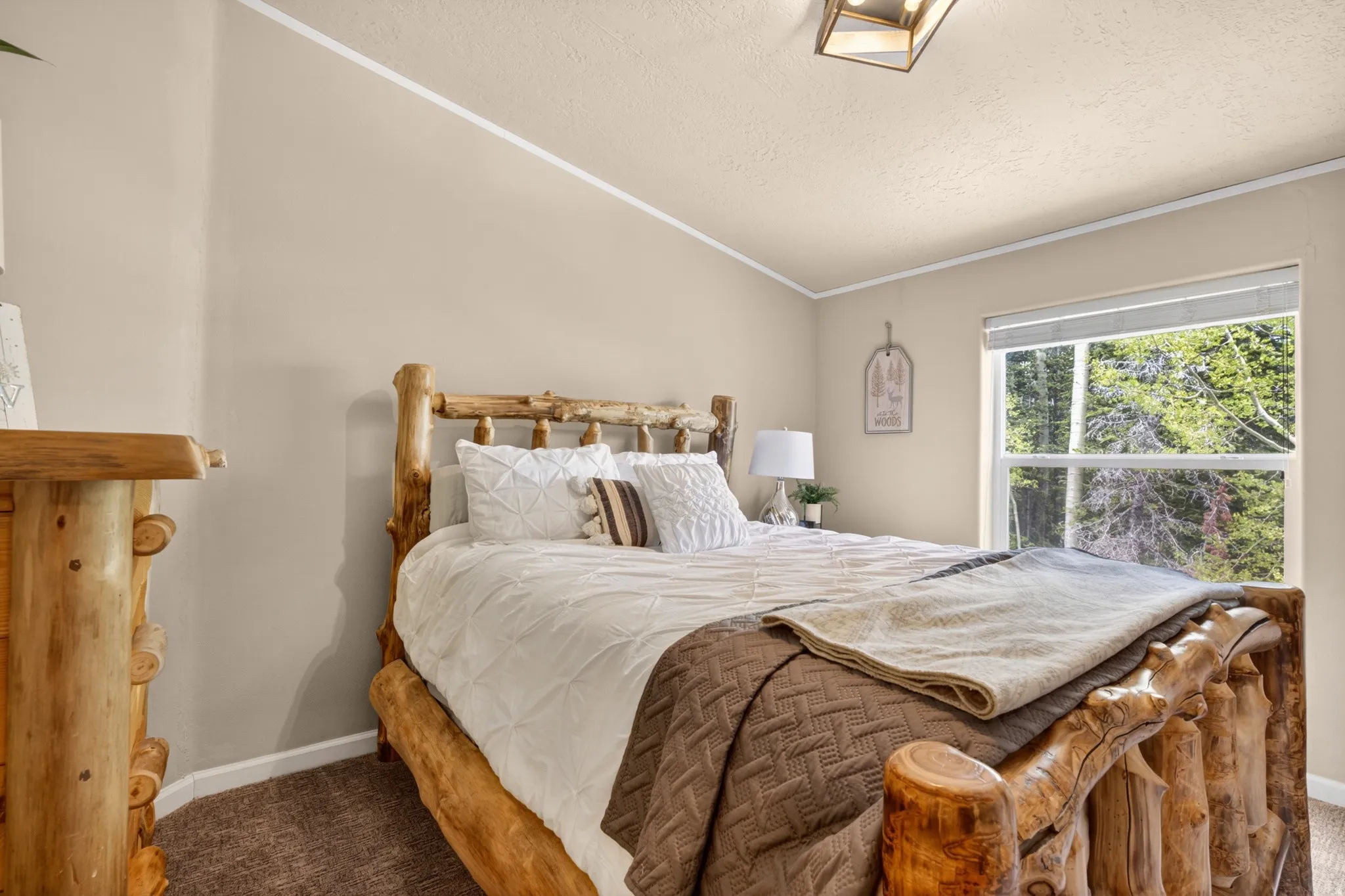 Spacious bedroom featuring natural wood bed frame and dresser, window with verdant tree scenery, neutral-toned walls, and recessed lighting
