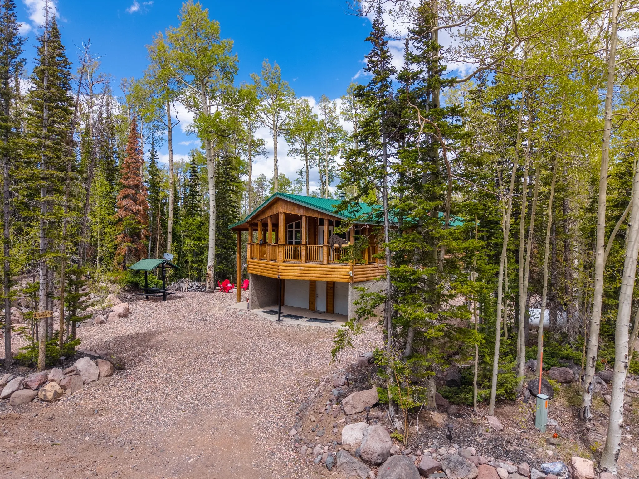 Log cabin exterior featuring a green metal roof, a covered balcony with wood railing, and a gravel driveway