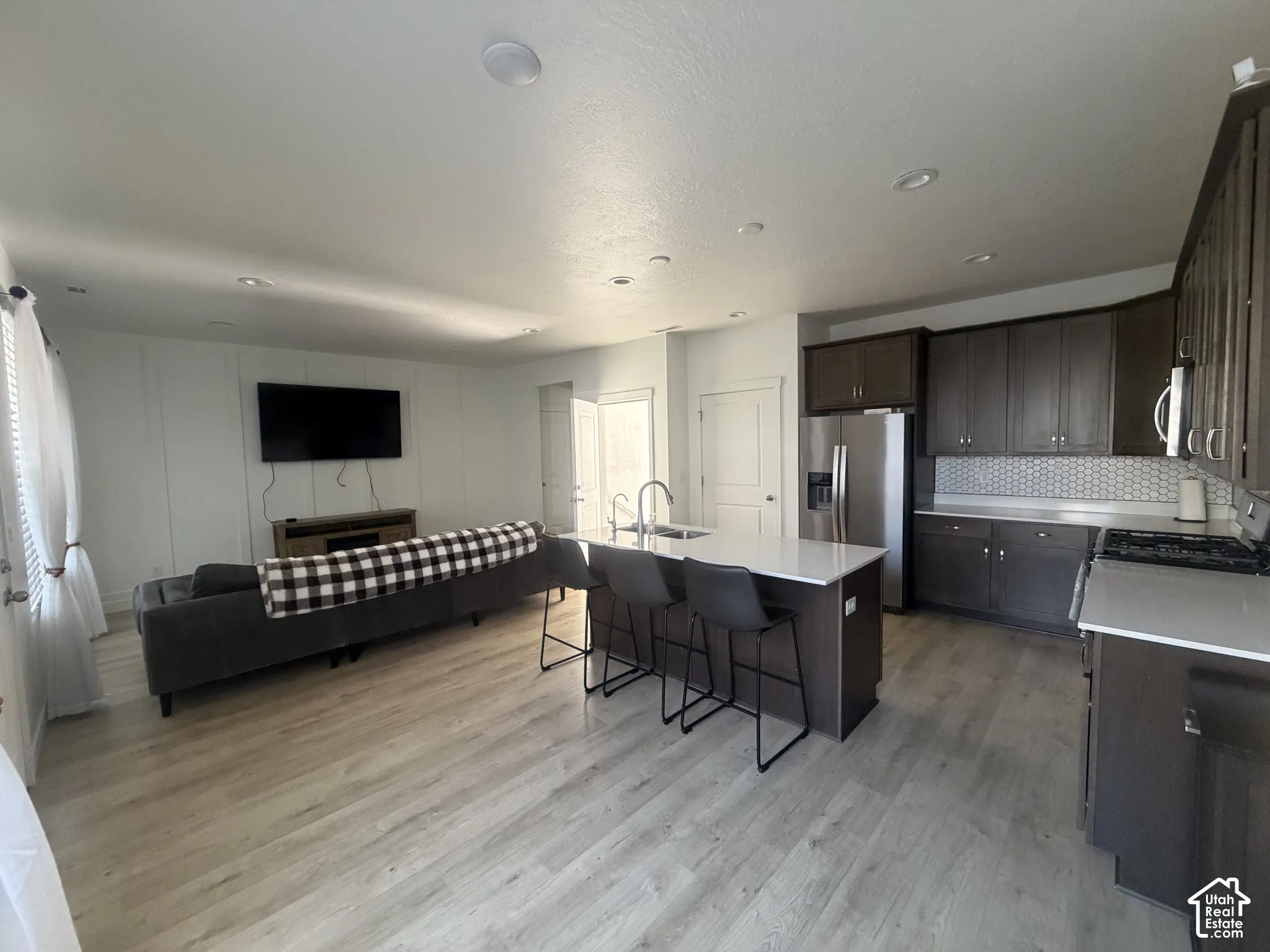 Open-concept interior featuring wood-finish flooring, dark-toned cabinetry, quartz countertops, a central kitchen island with a built-in sink, and hexagonal tile backsplash. complete view of living room and dining room