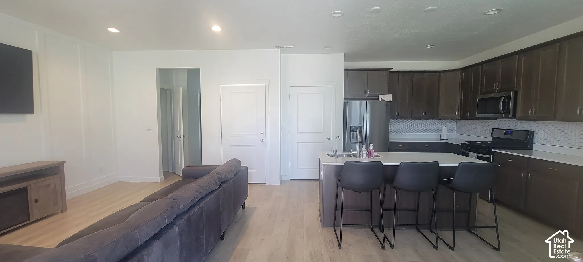 Open-concept living space featuring wood-finish flooring, recessed lighting, a kitchen island with seating, a patterned tile backsplash, and stainless steel appliances