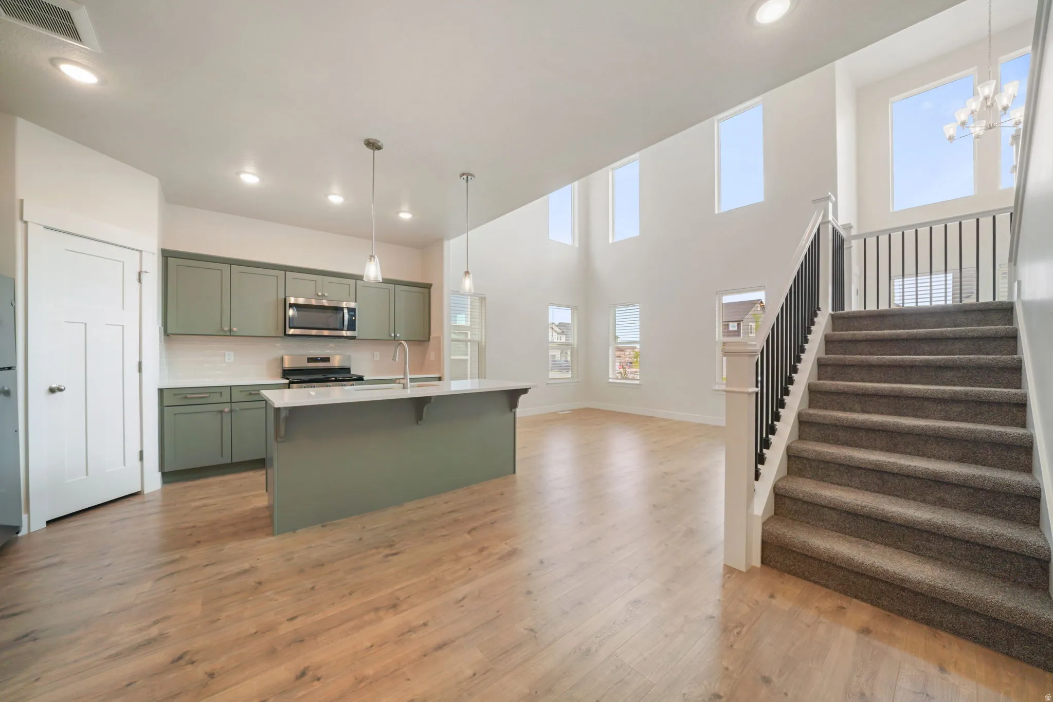 Open-concept living space with wood-finish flooring, a contemporary kitchen featuring sage cabinetry and white subway tile backsplash, and a central island with a sink and pendant lighting
