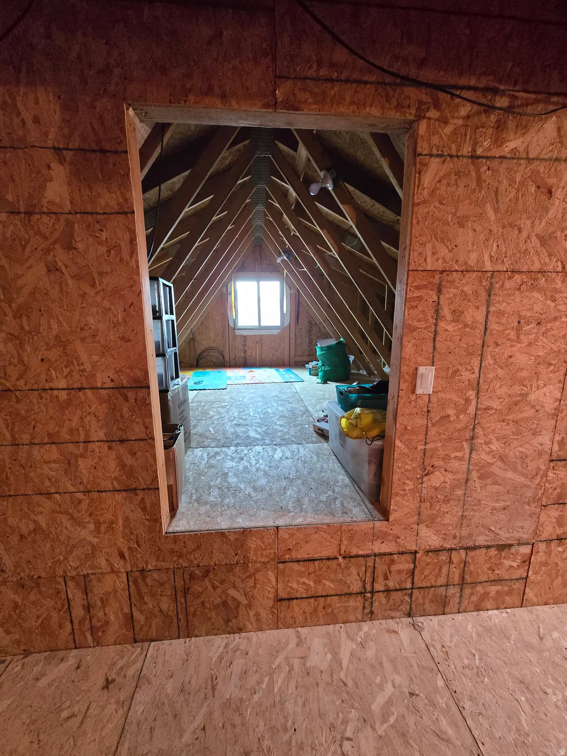 Attic space featuring exposed wood framing, a single window, and unfinished oriented strand board walls and flooring