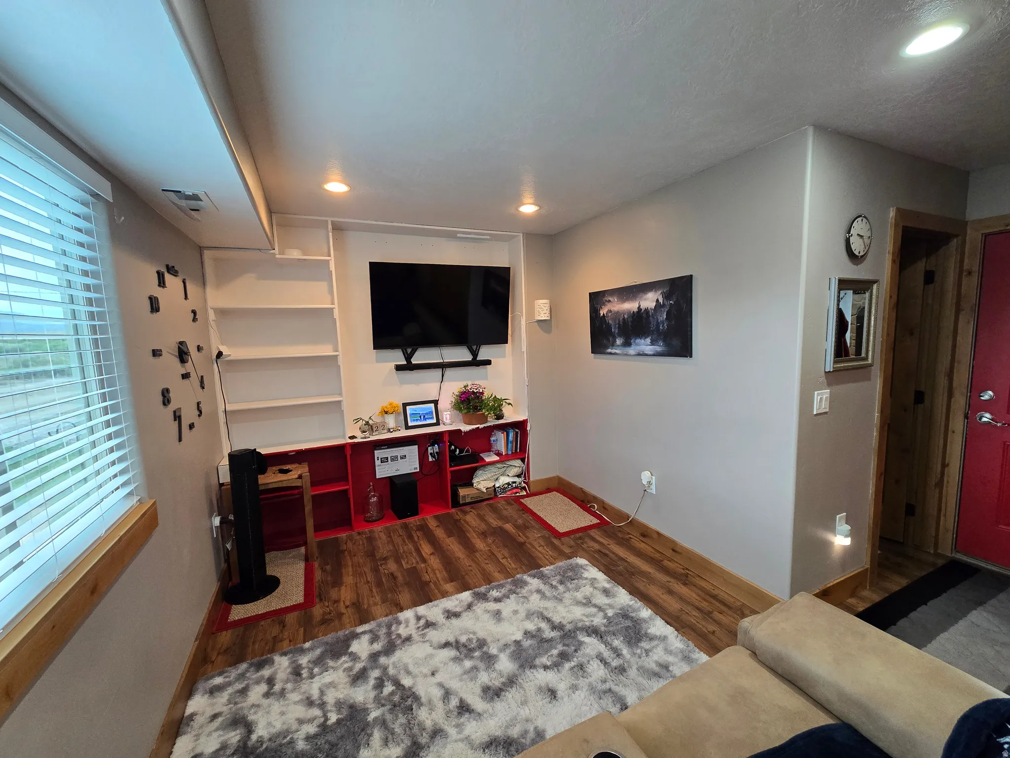 Living area featuring wood-finish flooring, recessed lighting, built-in shelving, a window with blinds, and a red interior door with brushed nickel hardware