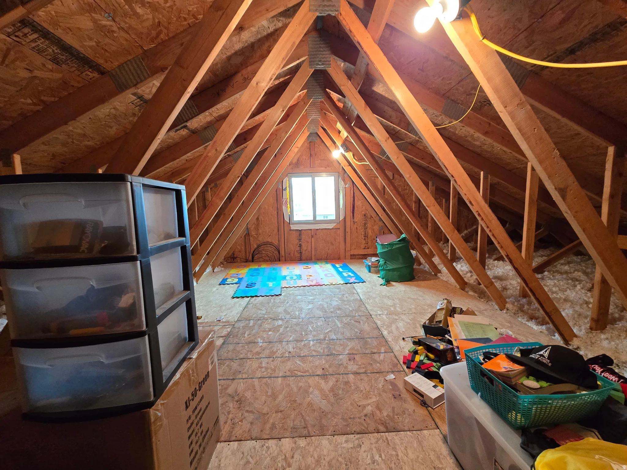 Spacious attic featuring exposed wood framing, plywood flooring, a single window, and visible insulation