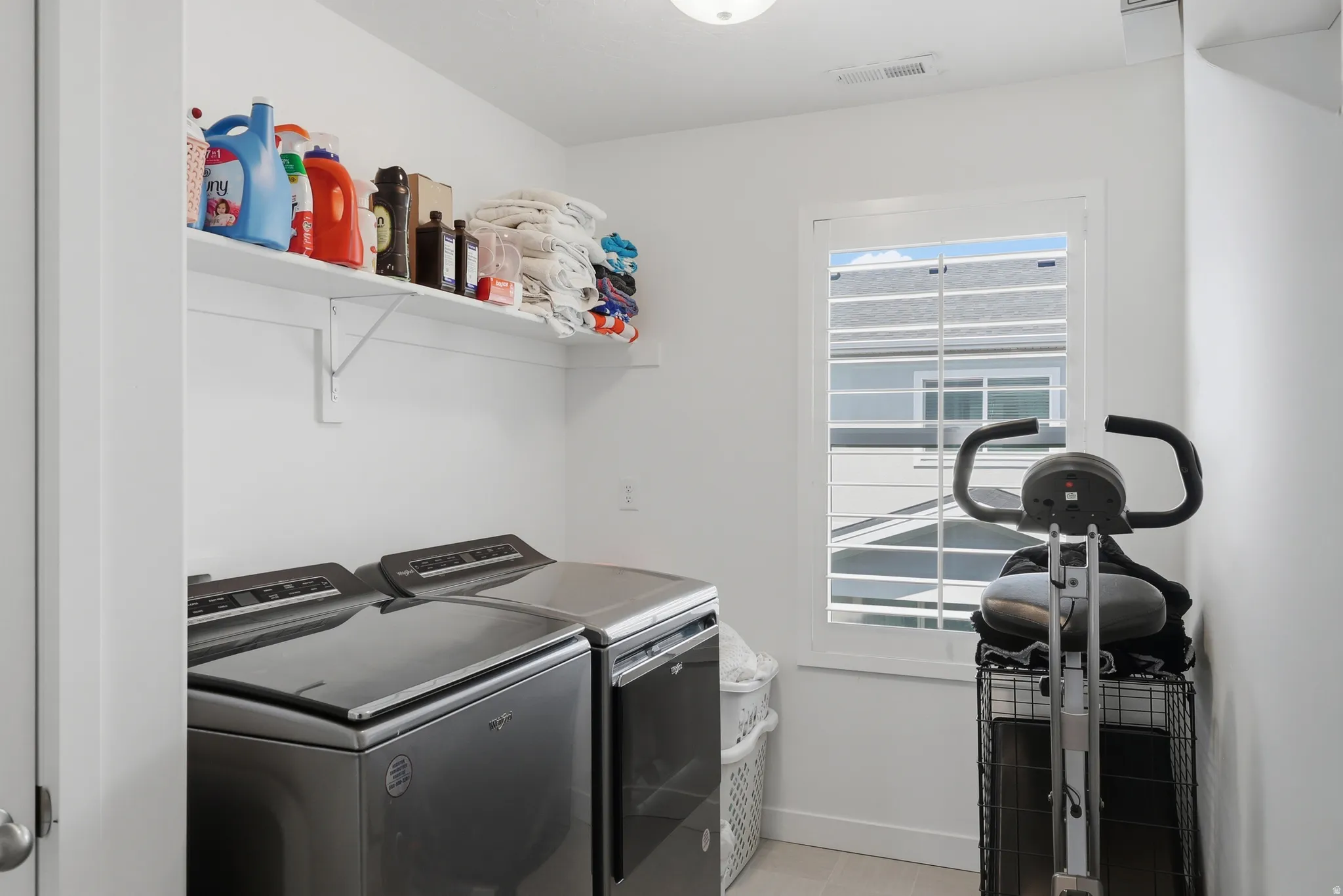 Bright utility space featuring a window with plantation shutters, overhead shelving, and light-toned flooring