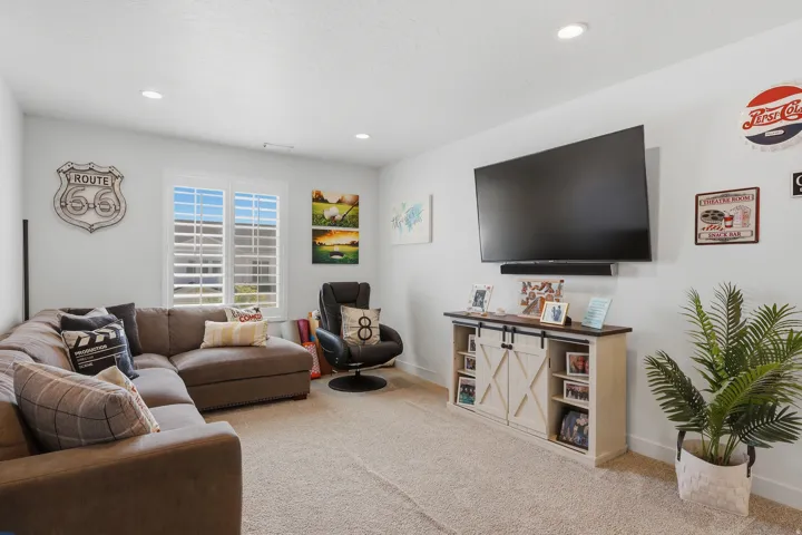 Carpeted entertainment area featuring a wall-mounted display, recessed lighting, and a window with plantation shutters