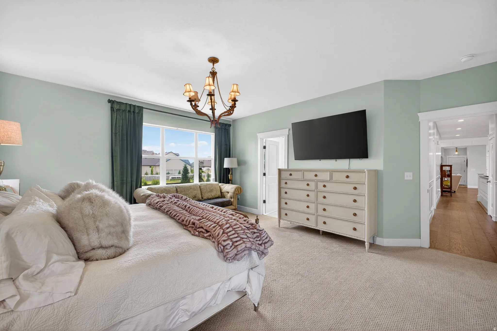 Spacious room with a large window, a classic chandelier, light green walls, light-colored carpeting, and a visible hallway with wood-finish flooring