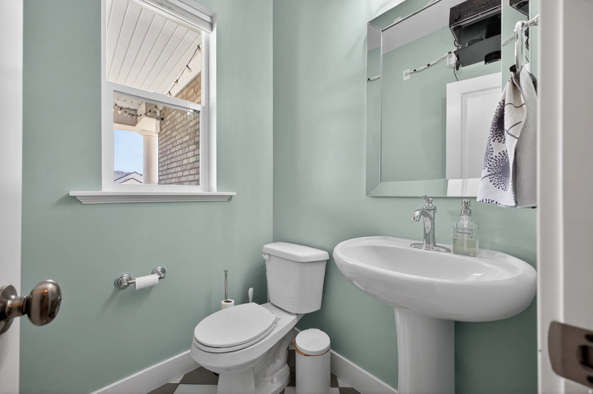 Pedestal sink with chrome faucet, white toilet, and window with a view of a brick exterior